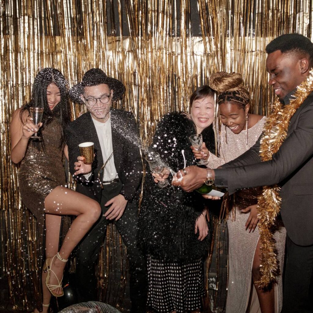 Group of people dressed up and having a great time with a gold tinsel backdrop and a bottle of champagne spraying