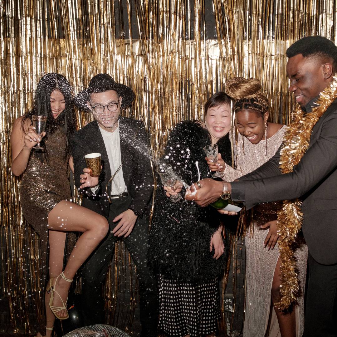 Group of people dressed up and having a great time with a gold tinsel backdrop and a bottle of champagne spraying