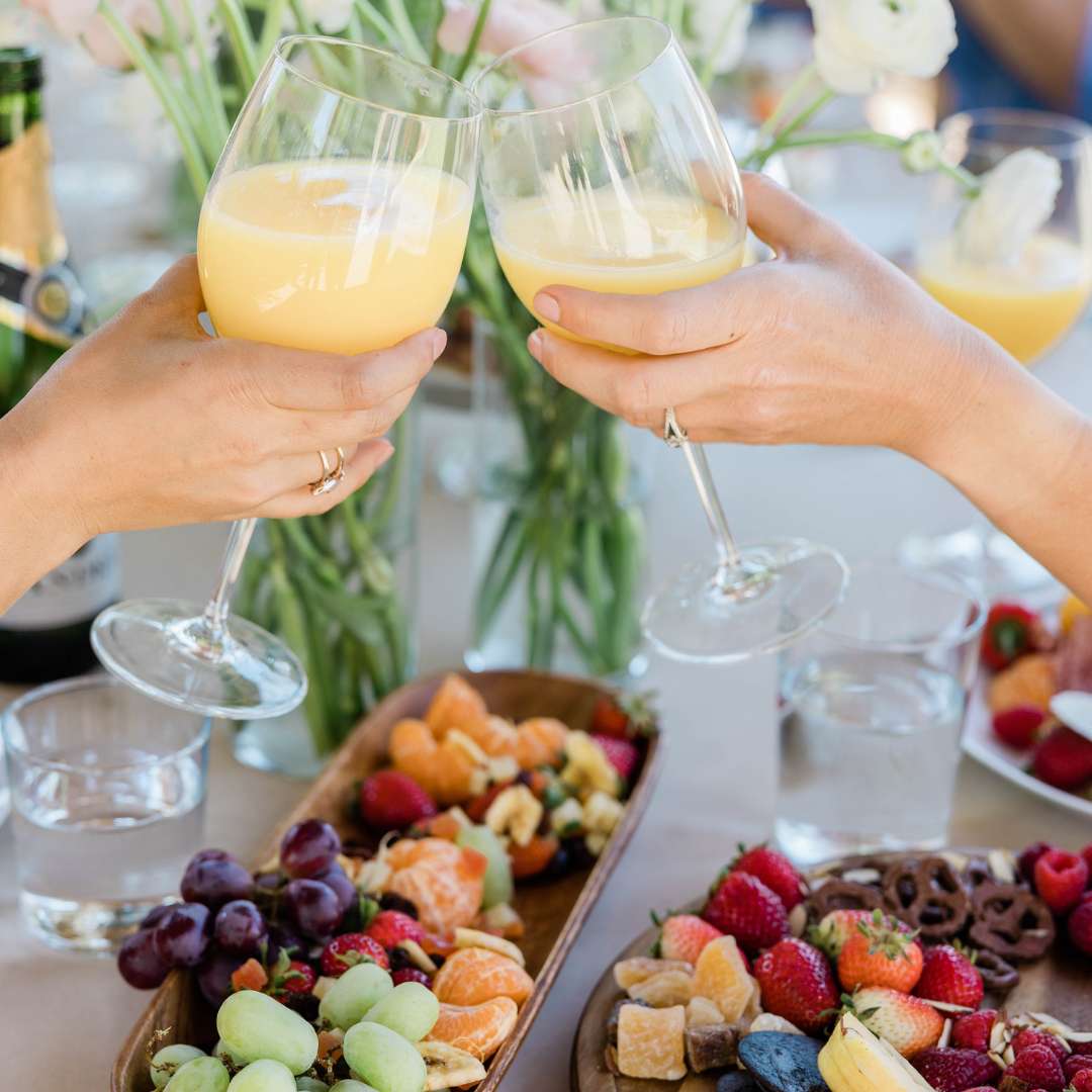 Two glasses full of mimosas toasting over a beautiful bowl of fruit and cheese